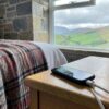 Prise Écosse : téléphone en charge sur une table de chevet dans une chambre de cottage écossais en pierre, plaid écossais et vue sur les Highlands