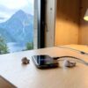 Prise Norvège : téléphone et écouteurs en charge sur un bureau en bois clair d'une cabane, fenêtre ouverte sur un fjord norvégien